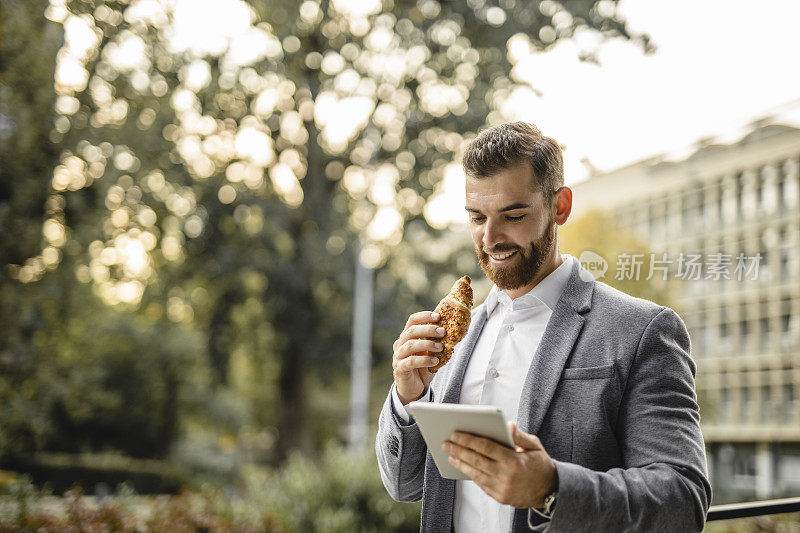 商人工作的肖像，使用数字平板电脑和吃牛角面包的户外