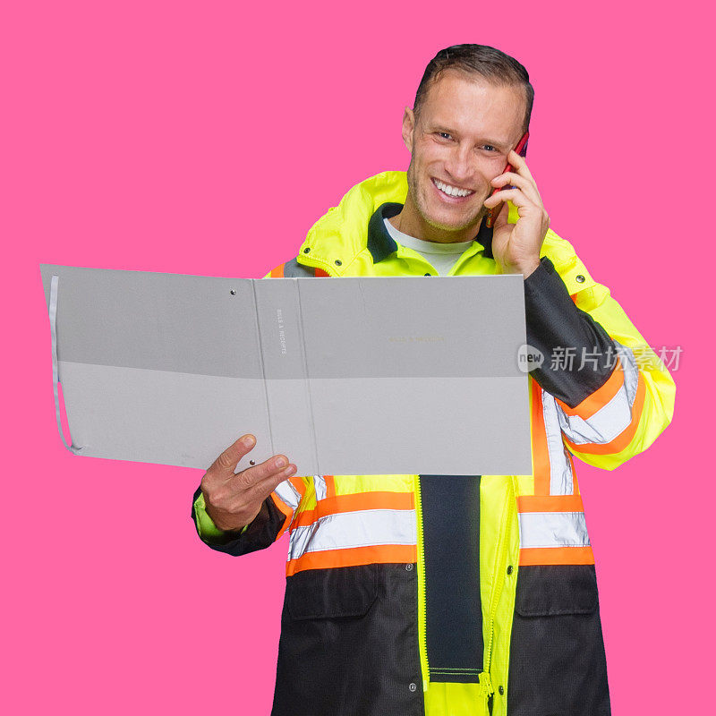 男性建筑承包商站在有色背景前，穿着背心，拿着活页夹，用手机