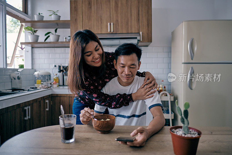 可爱的妻子在丈夫吃早餐时拥抱丈夫