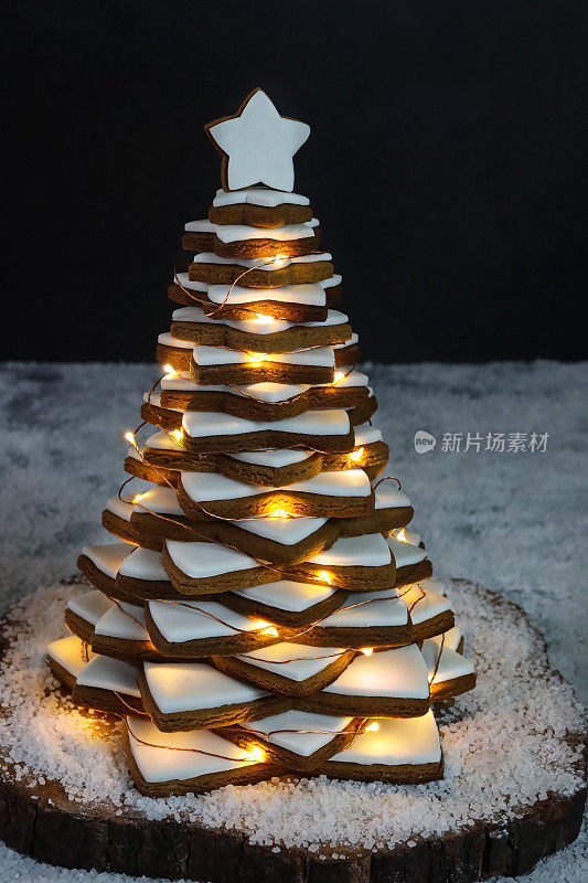 近距离拍摄的圣诞树是由堆在木蛋糕架上的姜饼星饼干做成的，上面覆盖着白色的皇家糖霜，被灯光照亮的仙女灯装饰着，周围是人造雪，黑色的背景，集中在前景上