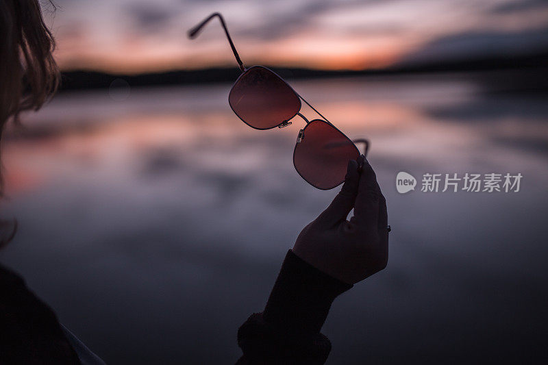 一个面目全非的女人站在湖岸，手里拿着太阳镜的特写