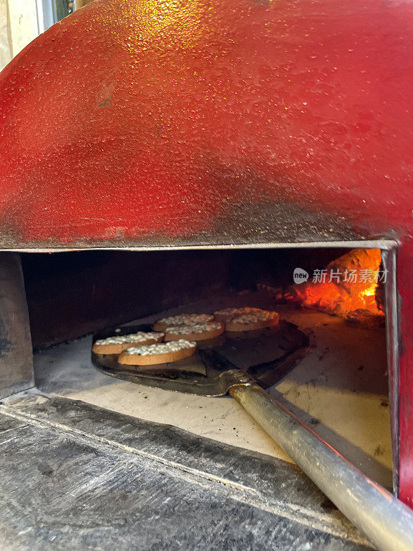 迷你披萨被滑入户外的形象，不锈钢，石基披萨烤箱长柄披萨桨，燃烧的木材，发光的火焰，重点在前景
