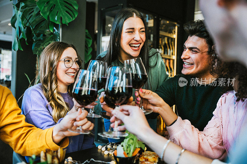 一群朋友碰杯欢呼，红酒杯坐在餐桌酒吧餐厅-年轻人在酒庄酒吧享受快乐时光-生活方式概念