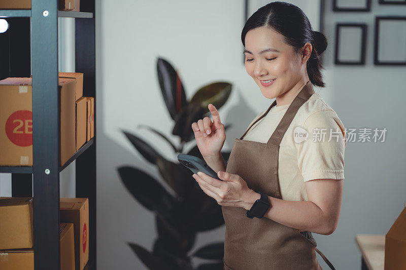 亚洲女性在线卖家使用智能手机，在家中办公室查看库存产品。