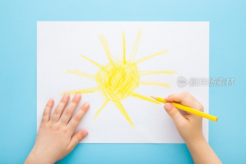 小孩子手拿黄色铅笔，在淡蓝色的桌子背景上白纸上画着大大的太阳。柔和的颜色。特写镜头。视角镜头。孩子创造时间。自顶向下视图。