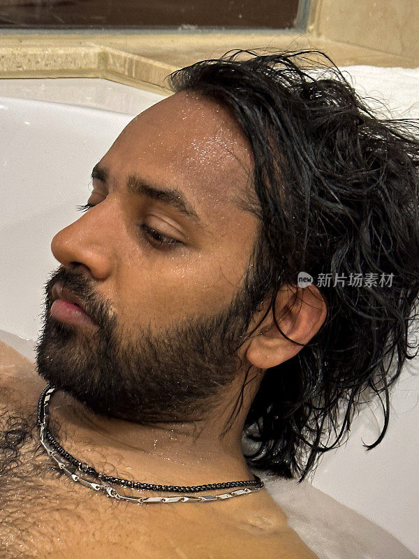 全画幅的印度男子躺在装满水的白色浴缸里，在浴缸里放松地把头靠在浴缸边的毛巾上，毛茸茸的胸部，面部毛发，头像，侧面视图