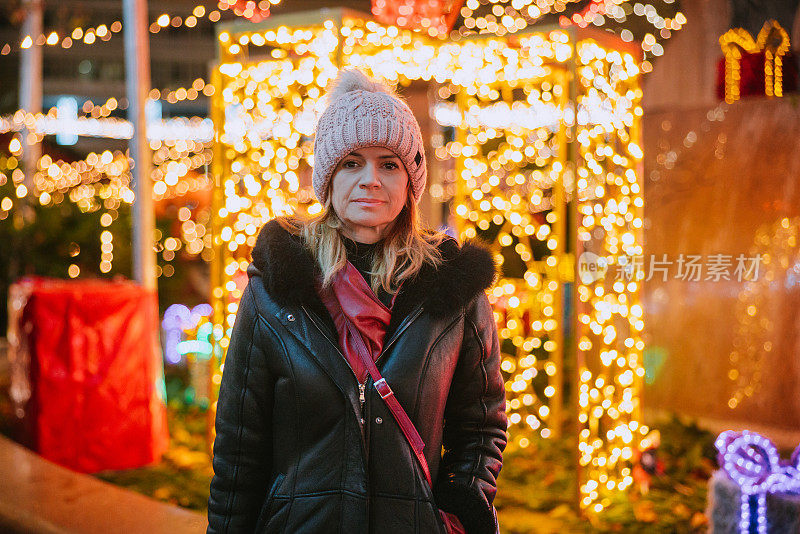 穿着冬季夹克的妇女站在装饰着彩灯的圣诞市场旁，看着镜头