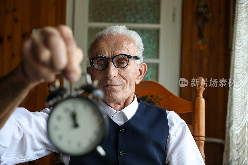 老人坐在扶手椅上，手里拿着一个小闹钟。