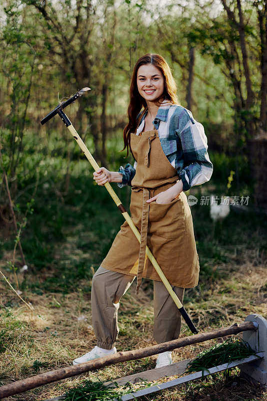 一个女人美丽地微笑着看着镜头，一个农民穿着工作服，系着围裙，在大自然中户外工作，拿着耙子为花园里的动物收集草和饲料