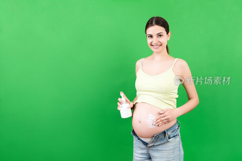 一名孕妇穿着敞开的牛仔裤，在彩色的背景上用面霜涂腹部的妊娠纹。孕期护肤概念