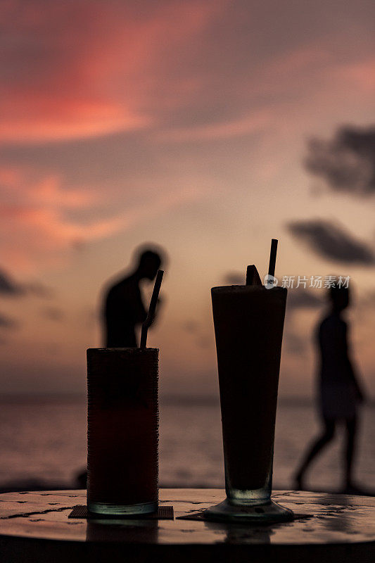 在一个热带岛屿上，两支鸡尾酒在日落时的剪影。人们从后面经过。快乐时光