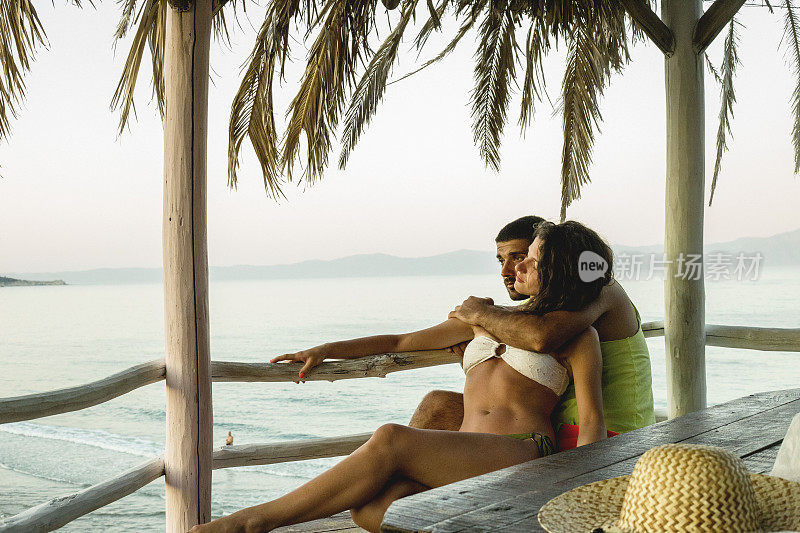 浪漫的夫妇坐在海滩上，享受美丽的海景，侧视图，花时间在一起，暑假的概念