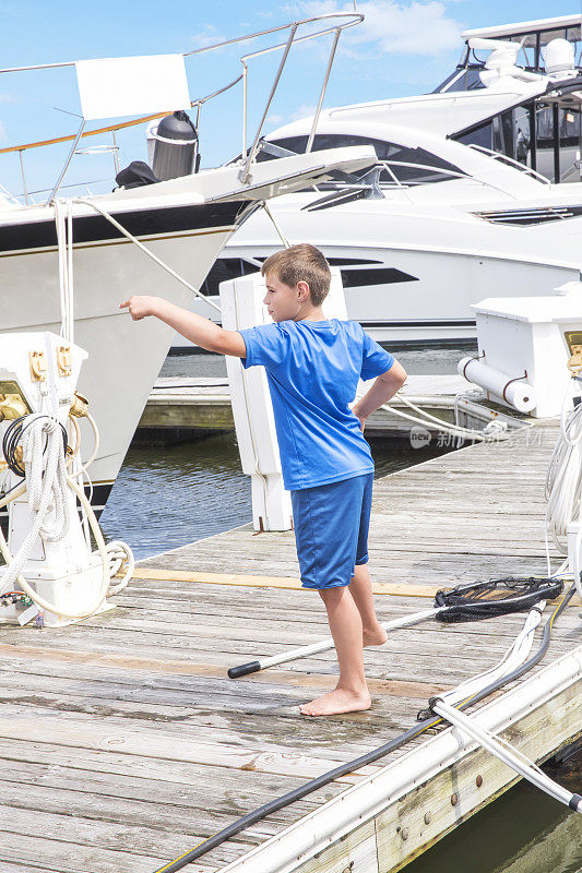 一个白人男孩站在他家游艇旁边的码头上