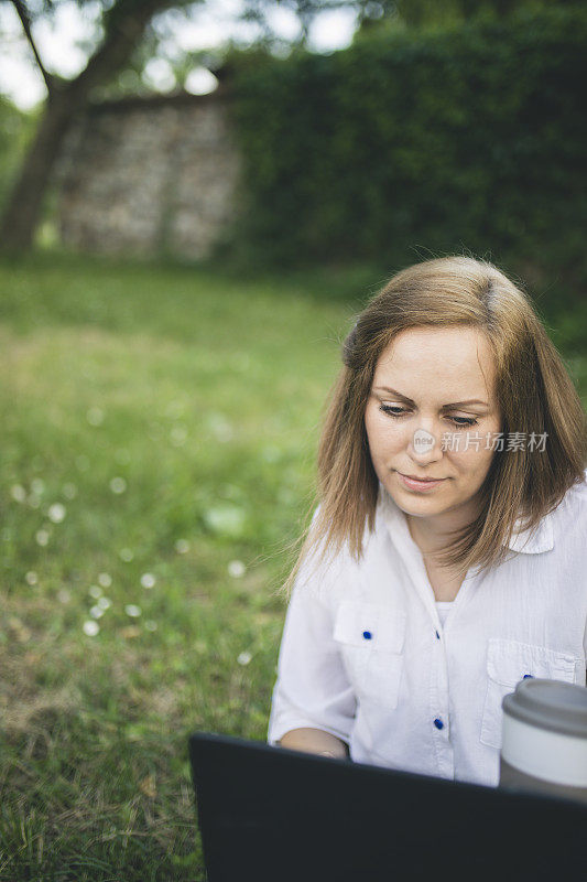 年轻迷人的女人拿着笔记本电脑和一杯咖啡或茶坐在草地上。