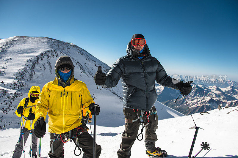 一名身穿羽绒服、背带的中年登山者在爬上一座白雪皑皑的山顶时，在他的朋友旁边竖起大拇指