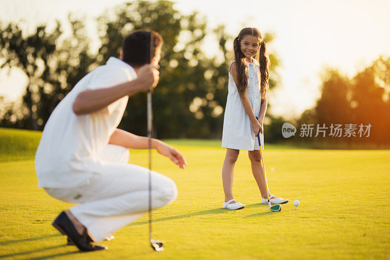 父亲蹲着，手里拿着一根高尔夫球棒，看着女儿，女儿看着他，准备击球