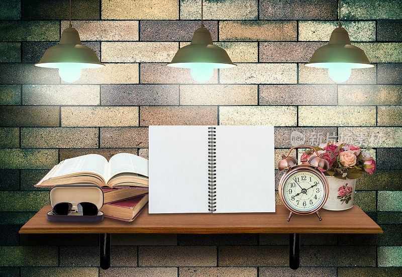顶部木质的架子上有空的上网本和打开的书，闹钟和玻璃在砖墙的背景上，以轻。产品展示概念