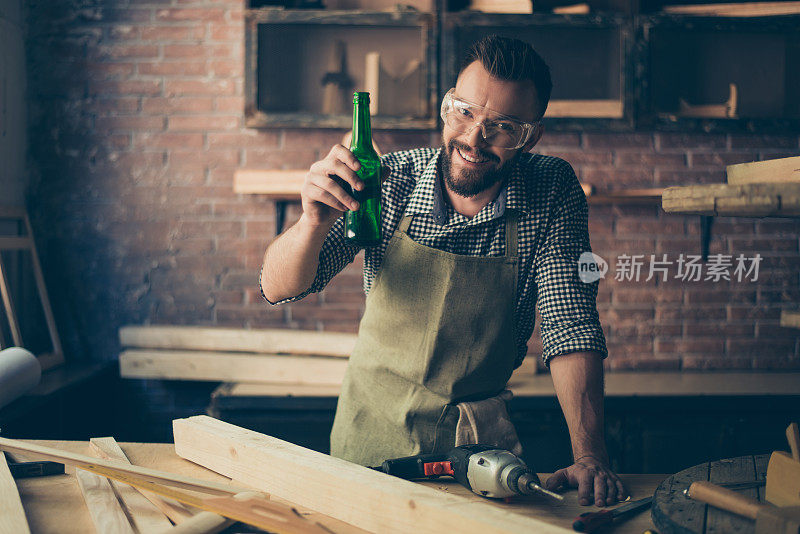 快乐愉快快乐放松的英俊胡子橱柜制造者是喝着啤酒庆祝结束一天的工作，他是站在书桌附近