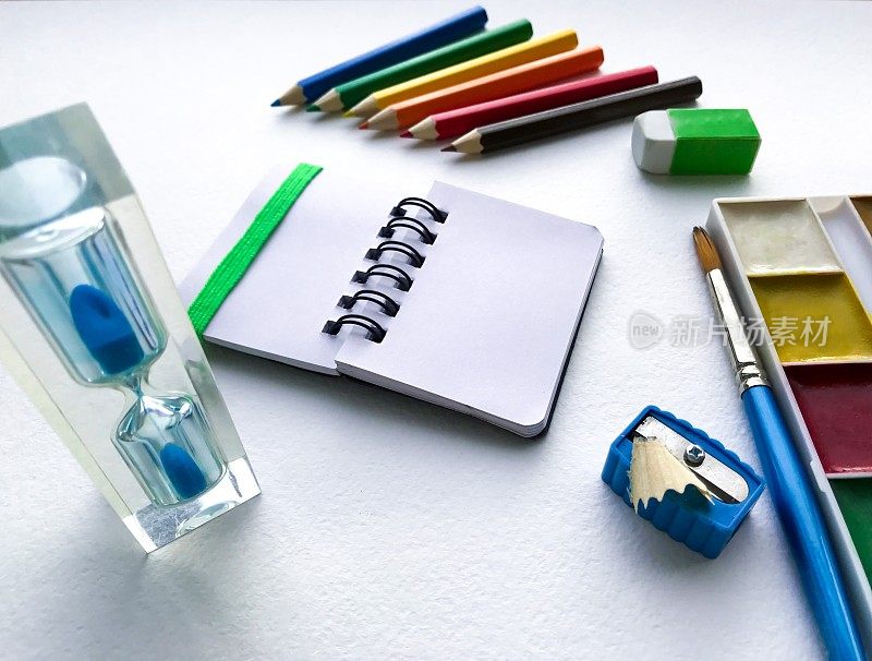 平铺文具学校和创意，绘画和工艺品(水彩画颜料，彩色铅笔，笔记本，沙漏)是在右边的白色水彩纸