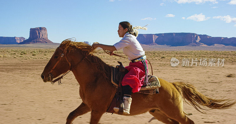 一个穿着传统纳瓦霍服装的十几岁美国土著女孩骑着她的马在一个晴朗、明亮的日子里穿越纪念碑谷沙漠和远处的大岩石群