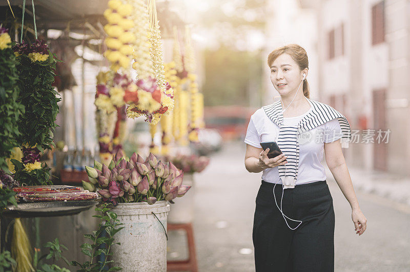 一位亚洲中国女游客在印度传统花店附近用耳机边听音乐边拿着手机走在街上