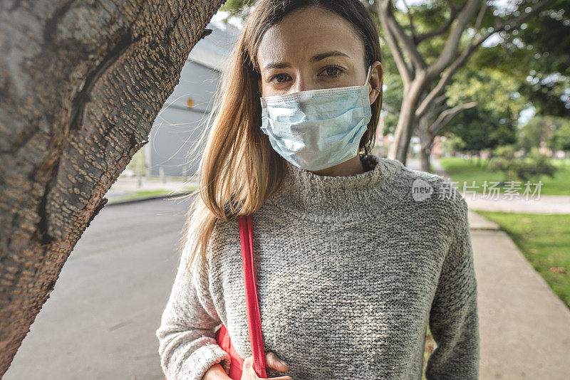 一名拉丁妇女走在街上，她戴着防护口罩，肩上背着一个用来把买回来的食物装回家的购物袋