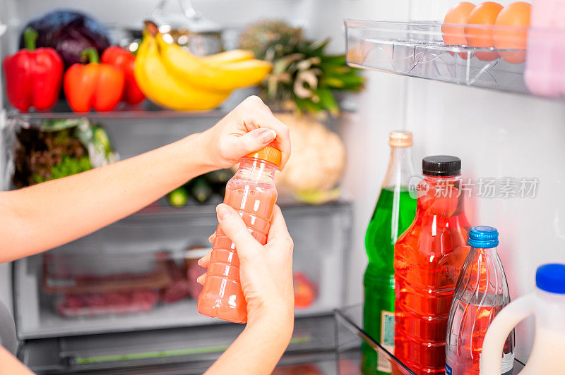 一个女孩打开一瓶粉红色的蔬菜或果汁或自制酸奶，背景是一个打开的冰箱和食物。饮食运动健康饮品概念，近景，浅景深。