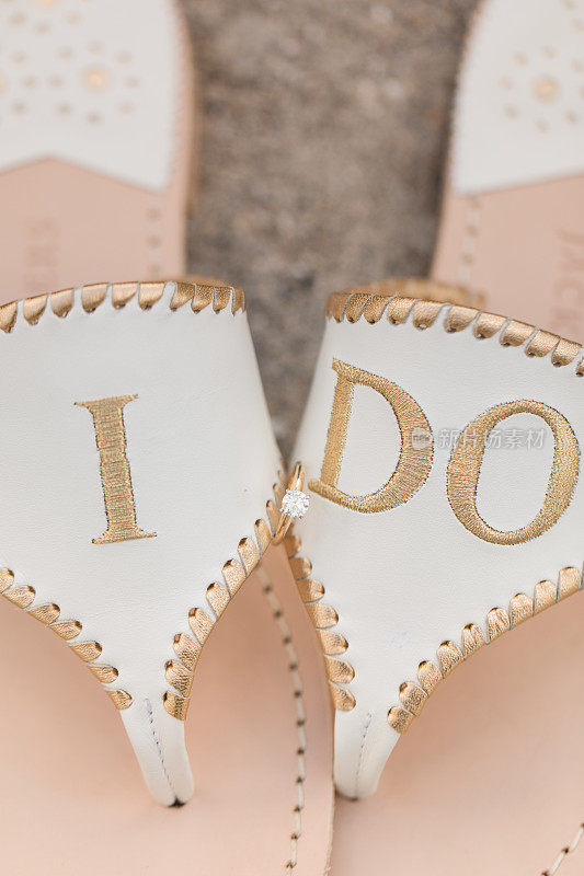 一双白色婚礼凉鞋上的钻石订婚戒指，上面的金字写着“我愿意”