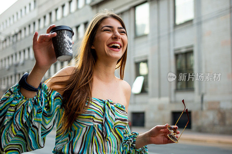 现代时尚的年轻女子在城市里享受着一杯咖啡，同时向朋友挥手