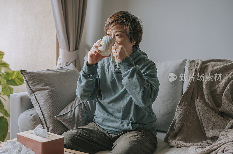 亚洲华人中年妇女喝着热茶不舒服，生病在家坐在沙发上休息看电视