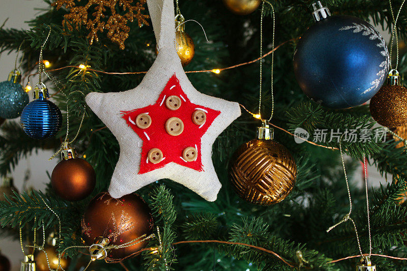 图像的毡和钮扣星装饰周围的蓝色，金色和铜彩色的小饰品挂在人造圣诞树树枝上，雪花，云杉针，模糊的白色仙女灯的背景