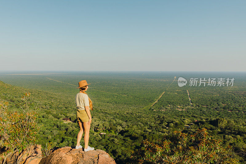 戴着帽子的女游客在纳米比亚的山顶上凝视着草原上的日落美景