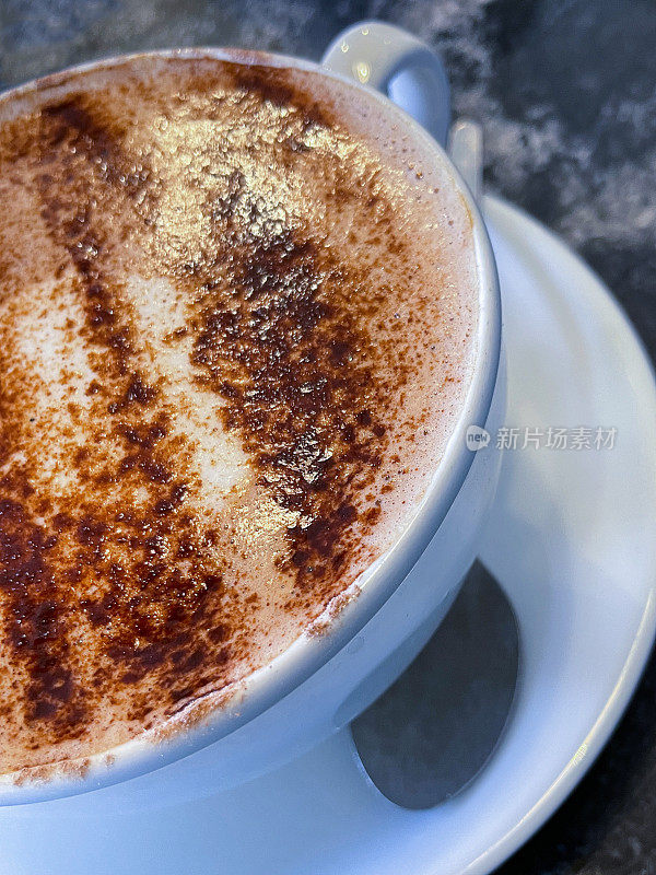 特写图像的咖啡桌场景，大的白色杯子和茶托与茶匙，热巧克力饮料，撒上可可粉，斑驳的黑色背景，高架视图