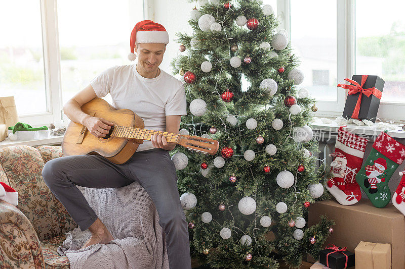 年轻英俊的男子坐在沙发上，他正弹着吉他，听着圣诞音乐，放松着概念