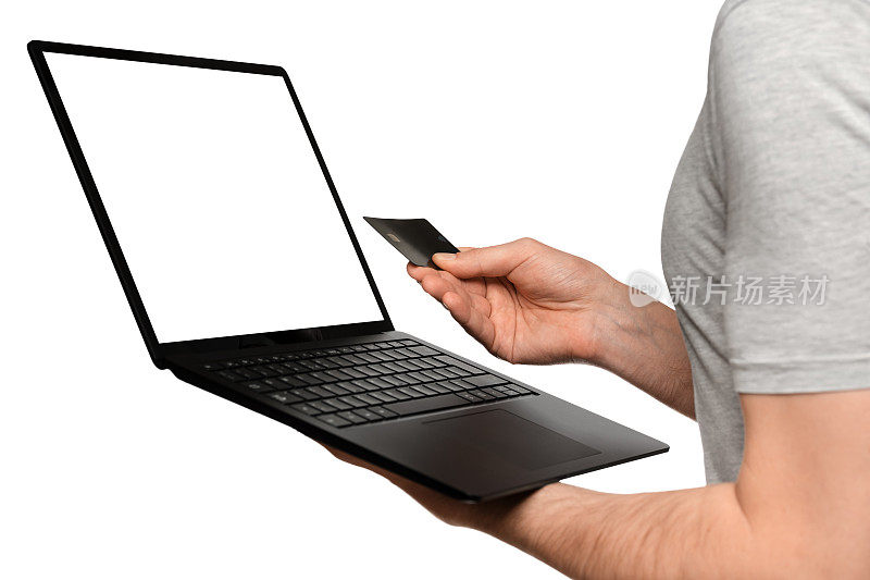 一名身穿t恤的男子手持一台笔记本电脑、一本超级本和一张银行卡，正在网上购物。