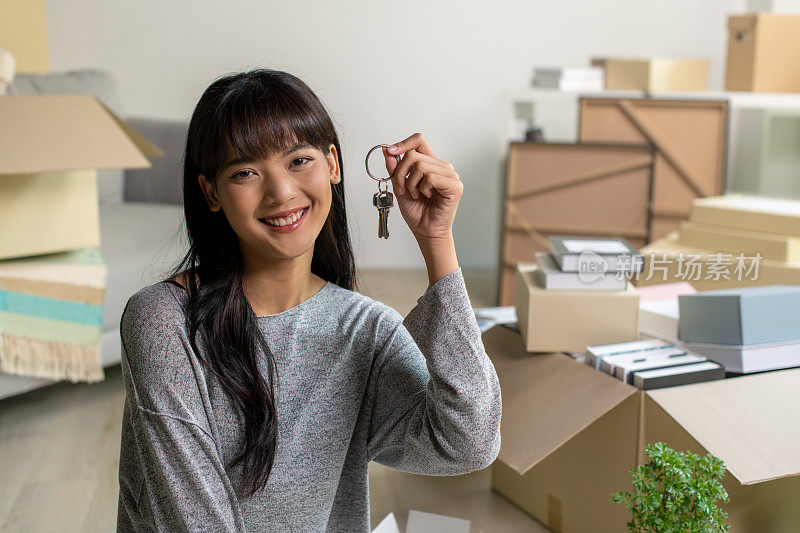 快乐的年轻亚洲女人拿着新房子的钥匙。周围都是纸箱和家具