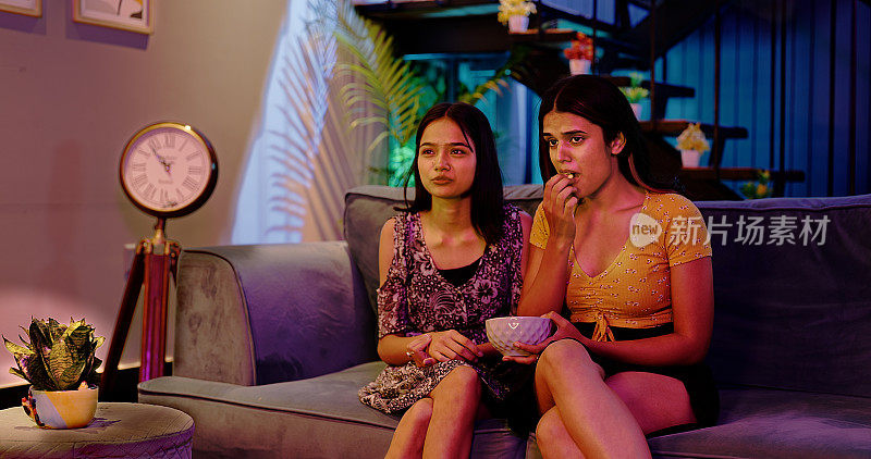 印度亚洲印度两个女人坐在沙发上看电视悲伤的电影一起吃爆米花零食在家里深夜。年轻漂亮的成年心烦女子擦着眼泪看着伤感的戏剧性真人秀度过周末时光