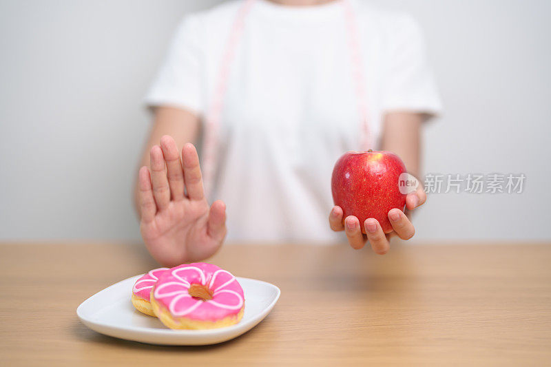 女性手持红苹果而拒绝甜甜圈，女性健身在健康水果和不健康的垃圾食品之间做出选择。节食控制，体重减轻，肥胖，饮食方式和营养