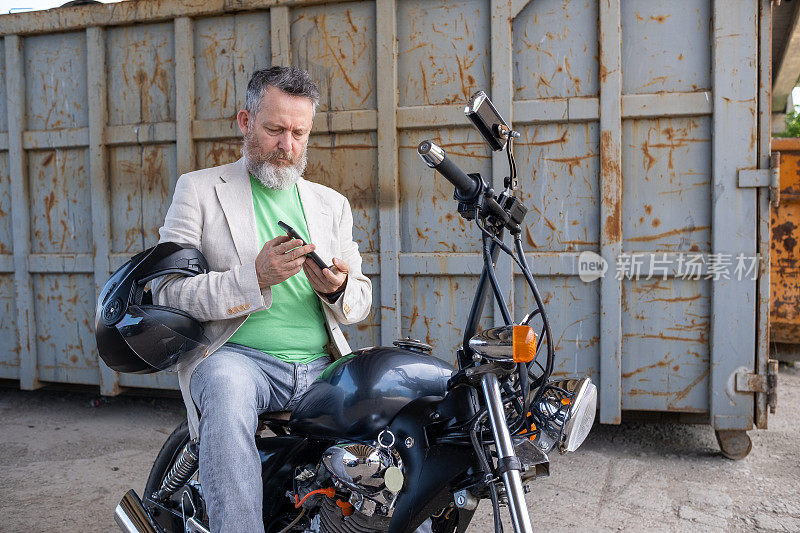 一个成熟的男人，穿着浅绿色的t恤，米色的夹克和灰色的牛仔裤，骑在一辆摩托车上。