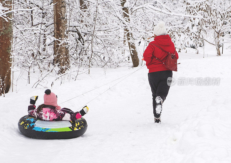 一名年轻女子跑着，抱着一个8岁的孩子，她骑在一个充气环上。一家人在下雪的公园里散步