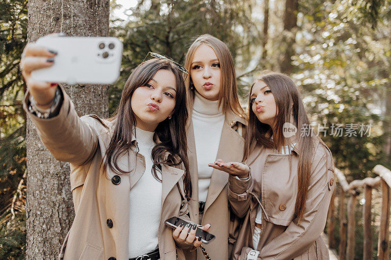 三位年轻女性在户外用智能手机自拍——快乐的美丽女性朋友在户外对着相机微笑——快乐女孩享受户外度假的生活方式概念。