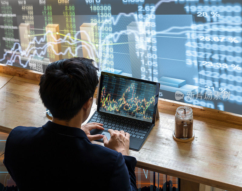 背面的商人坐在和使用电脑笔记本显示交易图形旁边的窗户玻璃上的股票市场图表背景，商业金融和交易概念
