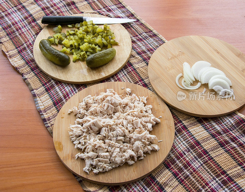 切碎的腌黄瓜放在砧板上，用刀切;木桌上放着煮熟的鸡肉和洋葱，上面放着格子厨房毛巾
