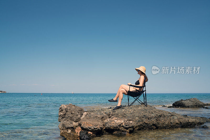 戴着帽子的年轻女子悠闲地坐在岩石岸边的椅子上