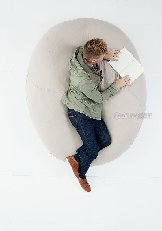 白种人年轻男学生躺在白色背景前，穿着暖和的衣服，拿着书