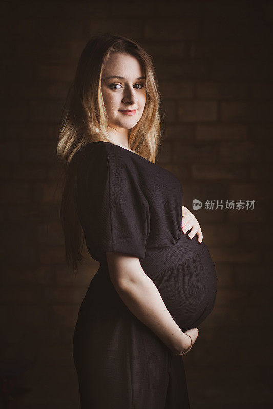 这是一幅以砖墙为背景拍摄的快乐、漂亮、年轻的怀孕妈妈的肖像，她有着金色的长发。