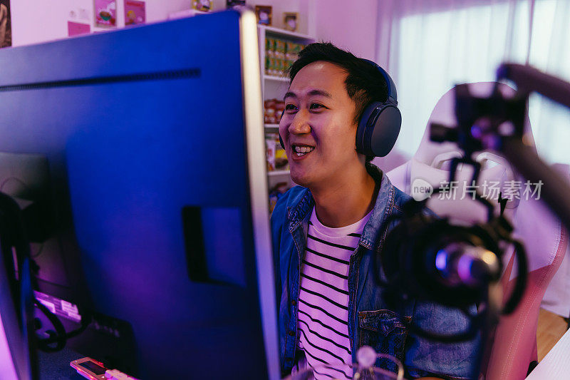 兴奋和微笑的男性玩家戴着耳机麦克风玩在线视频游戏。年轻的亚洲人在家里用个人电脑与玩家和观众交谈