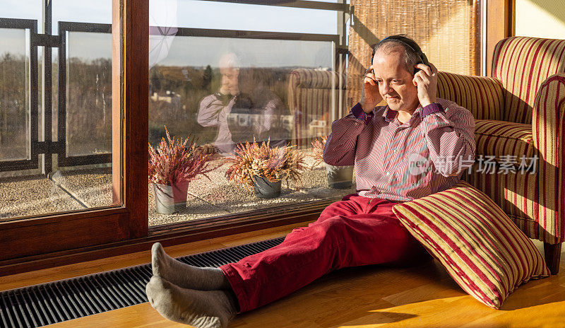 一位中年白人老人戴着耳机坐在家里，靠在有靠垫的扶手椅上听着音乐