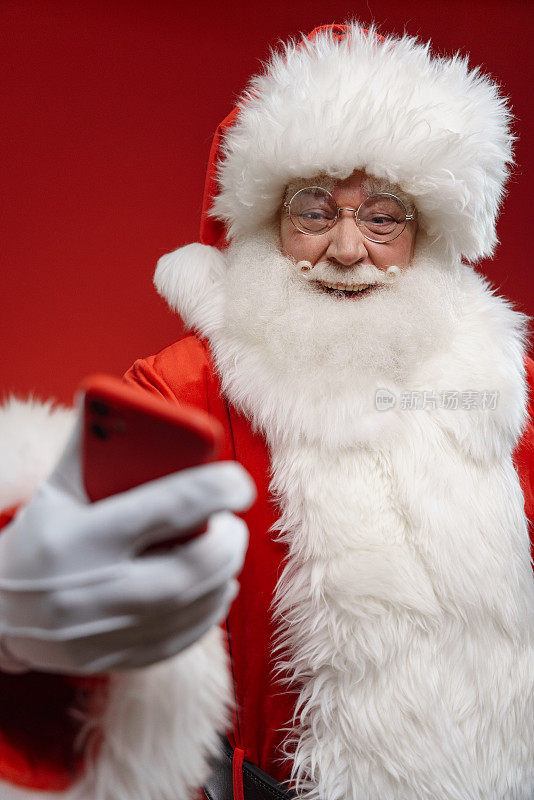 在摄影棚里，戴着眼镜的圣诞老人在明亮的红色背景上看着他的红色智能手机