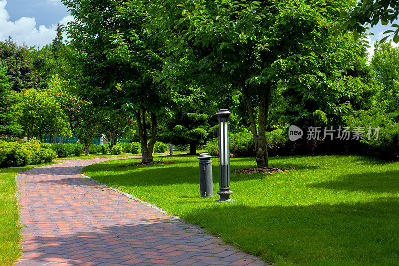 铁灯笼地面花园照明的人行道铺着石砖在公园的绿色植物，灌木和树木与覆盖在绿色草坪在一个阳光灿烂的夏天，没有人。